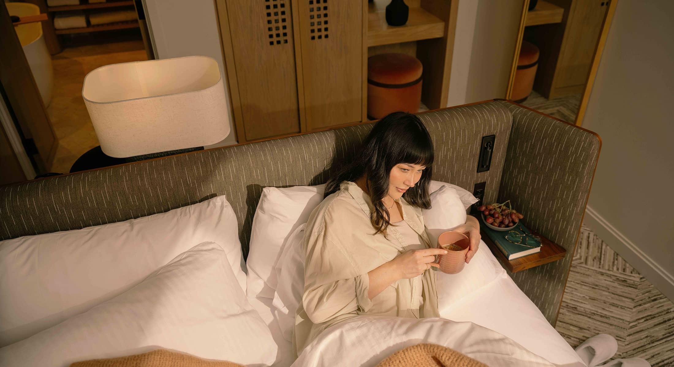 Woman relaxing in a Rimrock Banff suite bed, holding a warm drink, surrounded by soft white linens, natural wood details, and cozy neutral textures in warm morning light.