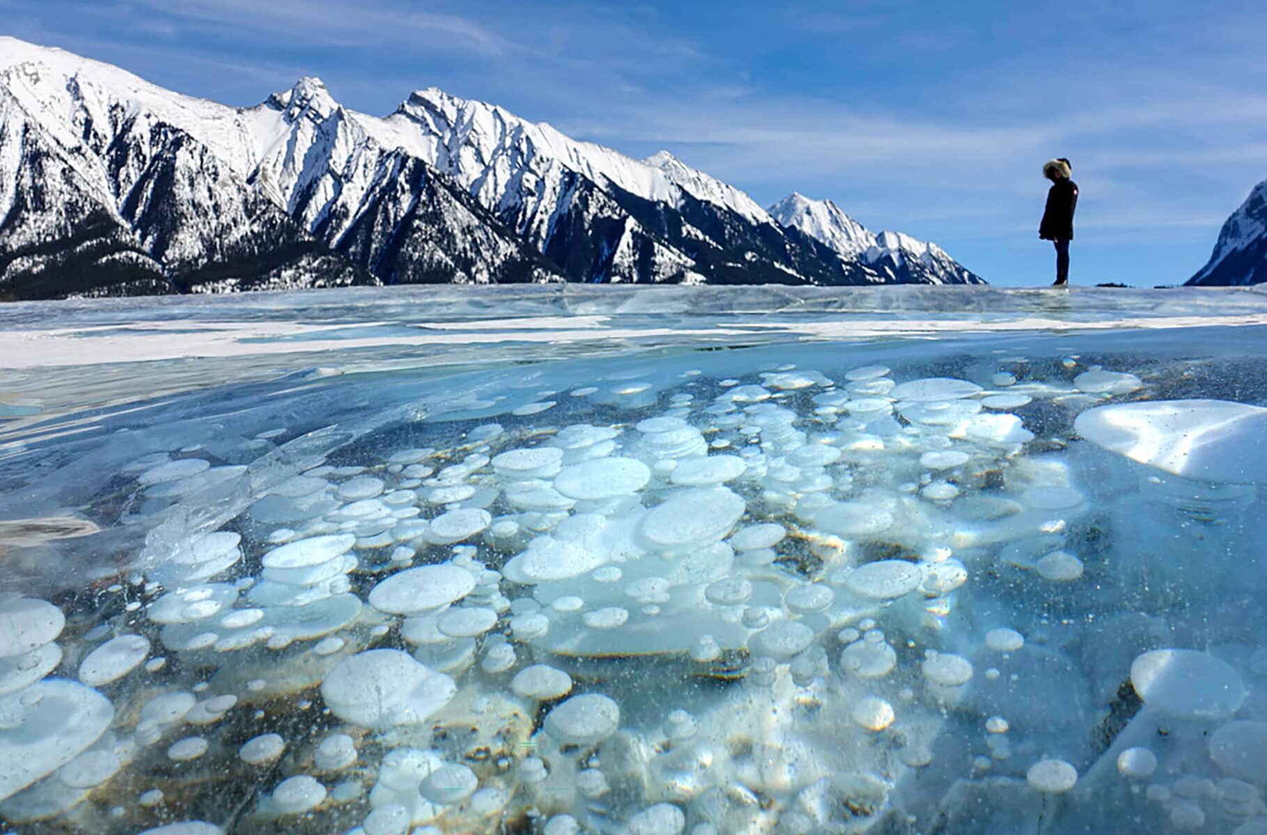 A person standing on the frozen surface of Abraham Lake, with layered ice bubbles visible beneath clear winter ice and mountains in the distance.