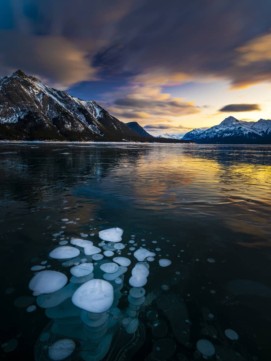 Methane ice bubbles trapped beneath the frozen surface of Abraham Lake at sunset, with surrounding mountain peaks reflected in the ice.