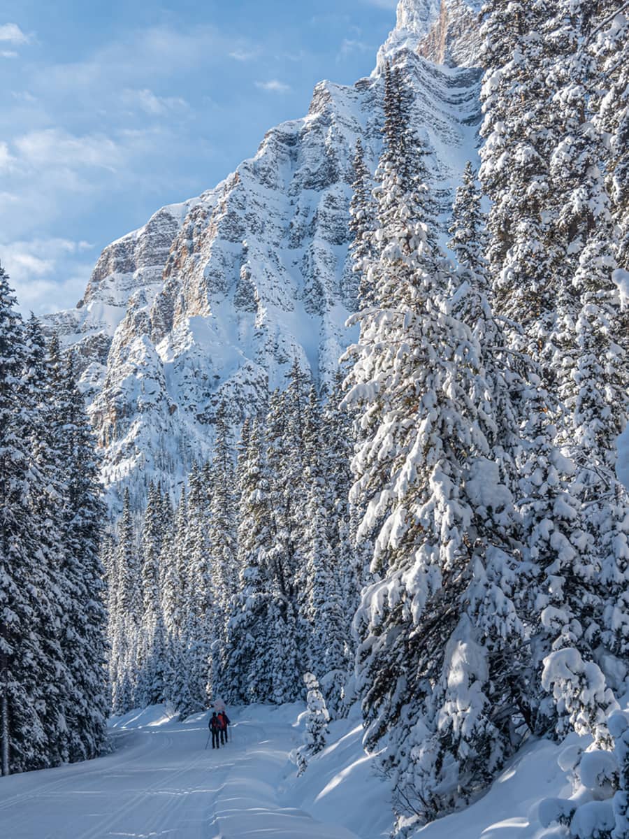 Two people snowshoeing together along a wide winter trail beneath steep, snow-covered mountain walls in Banff National Park.