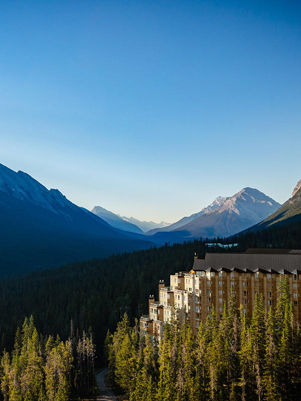 Aerial view of Rimrock Banff perched along Sulphur Mountain, surrounded by dense evergreen forest with sweeping views of the Bow Valley and Rocky Mountain peaks under a clear blue sky.
