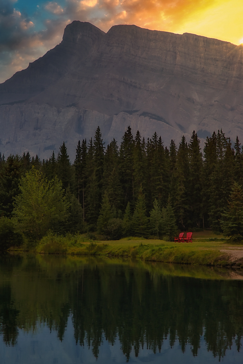 A bright sunny sky glowing over mountains at Cascade Ponds in Banff with red Parks Canada chairs on the shore.
