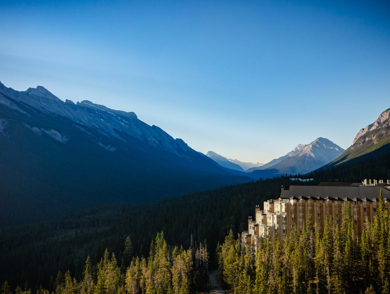 Aerial view of Rimrock Banff perched along Sulphur Mountain, surrounded by dense evergreen forest with sweeping views of the Bow Valley and Rocky Mountain peaks under a clear blue sky.