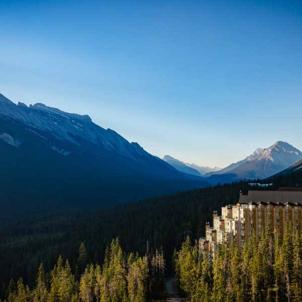 Aerial view of Rimrock Banff perched along Sulphur Mountain, surrounded by dense evergreen forest with sweeping views of the Bow Valley and Rocky Mountain peaks under a clear blue sky.