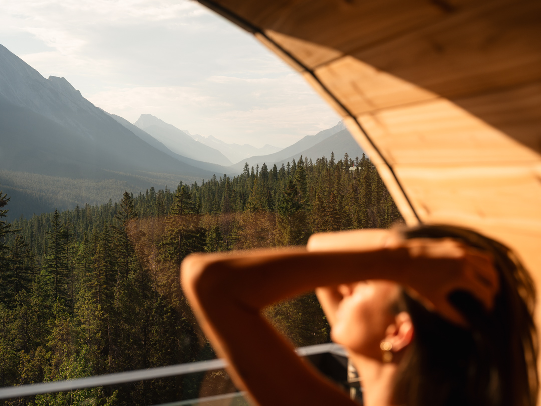 A woman with her fingers in her hair inside of the sauna at Rimrock Banff overlooking Spray Valley.