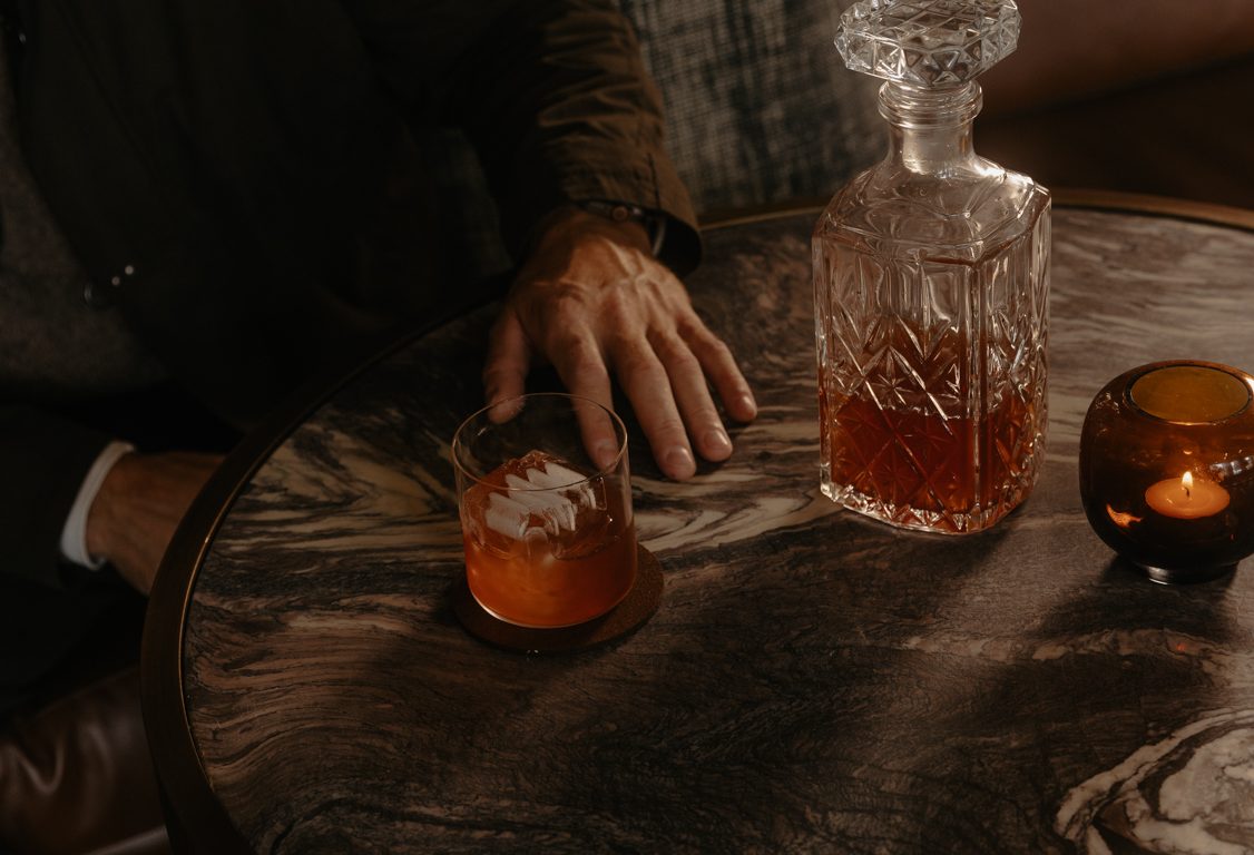 Guest seated beside a cocktail, crystal decanter and candle on a marble table in Listening Room at Rimrock Banff.