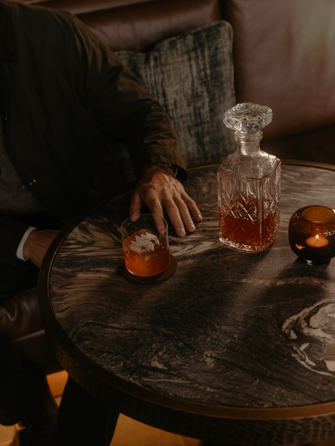 Guest seated beside a cocktail, crystal decanter and candle on a marble table in Listening Room at Rimrock Banff.