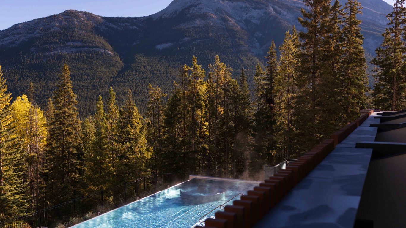 View from a Wellness Suite at Rimrock Banff overlooking the outdoor infinity pool, evergreen forest and Canadian Rockies.